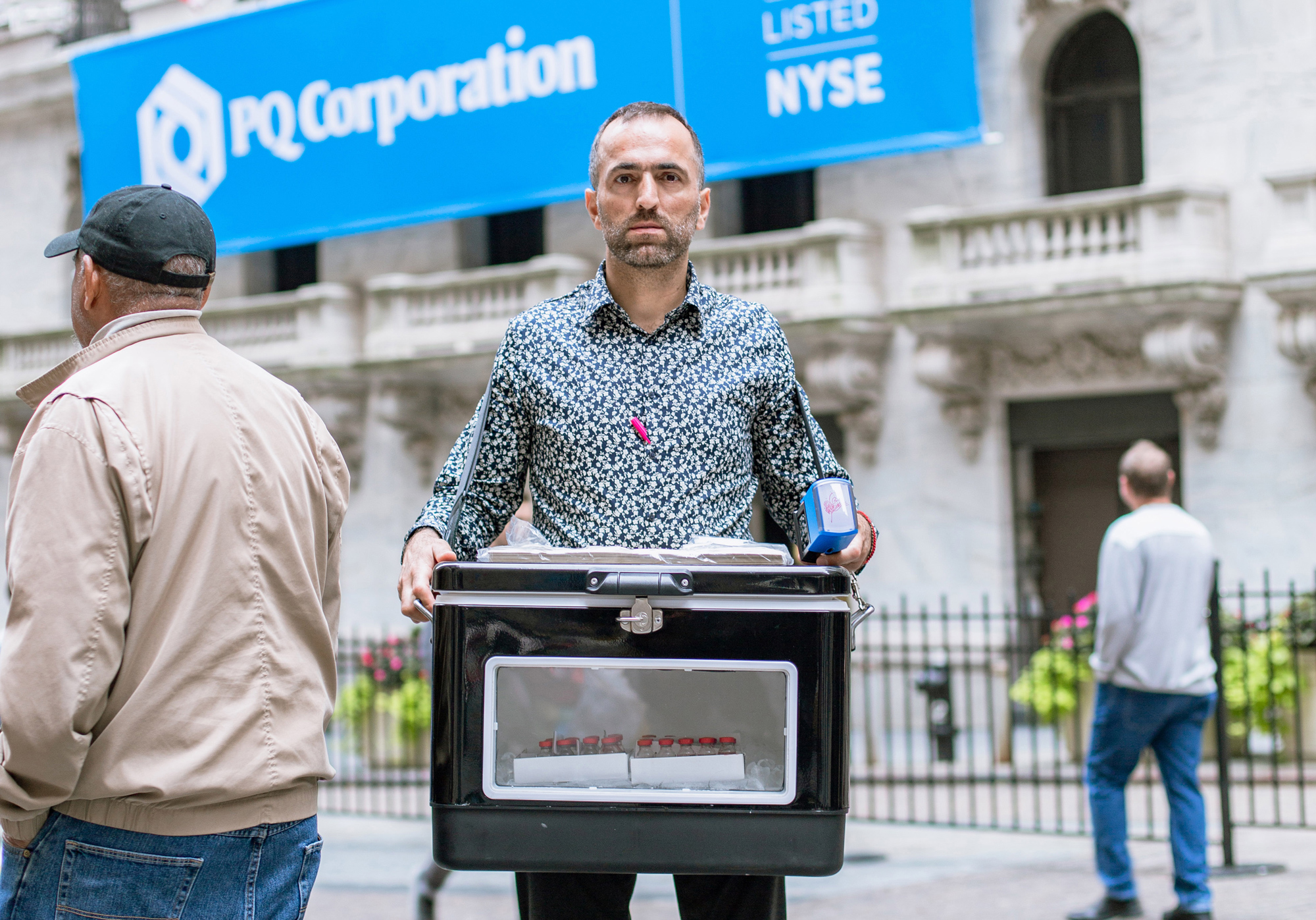 Image of Khaled Jarrar’s public performance on Wall Street. A man holds a black metal cooler. Glass viles can be seen through an opening in the front.