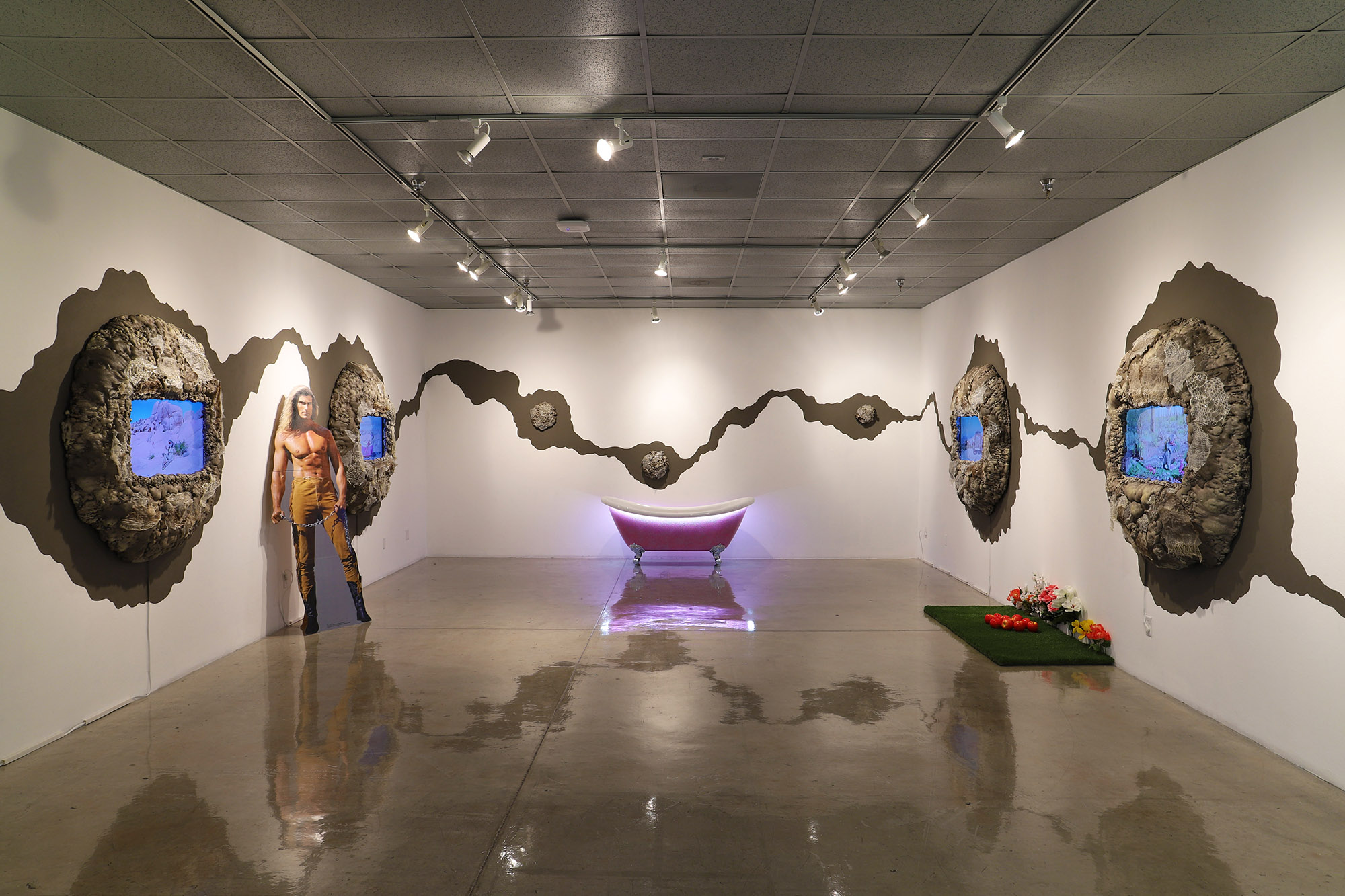 Image of Maria Shaltout’s Snake Woman multimedia installation. Four video screens set within brown mounds of dirt are hung on opposing white walls with brown painted uneven lines that surround the room and connect them all together. A pink bathtub with lights sits against the back wall and a life-sized cutout figure of a shirtless man stands against the left wall. Green turf and bunches of multi-colored silk flowers sit on the floor against the right wall.