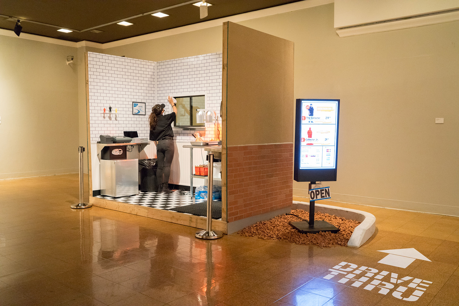 Image of Oliver Padilla’s Product of the American Dream installation. A small fast-food restaurant drive-thru with a three-sided white tile kitchen is constructed in the gallery. A person in a black restaurant uniform wipes down the tiled walls of the room.
